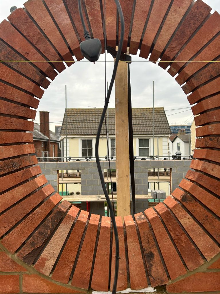 View through a circular brick arch highlighting construction work in the background, featuring a wooden support and electrical wiring.